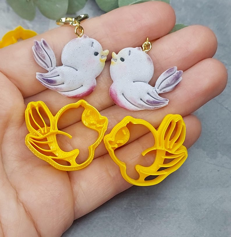 May include: A pair of white bird earrings with purple accents and a pair of yellow bird-shaped cookie cutters.