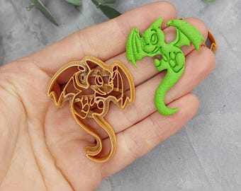 Könnte beinhalten: Ein goldfarbener Drachen-Ausstecher neben einem grünen Drachen-Keks. Die Ausstecher haben die Form eines Cartoon-Drachen mit Flügeln und einem langen Schwanz. Der grüne Drache ist ein fertiges Produkt.