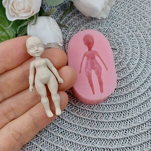 Op de afbeelding: Een roze siliconen mal voor het maken van een poppenfiguur. De mal is gevormd als een persoon met armen, benen en een hoofd. Naast de mal is een klein, wit poppenfiguur te zien.