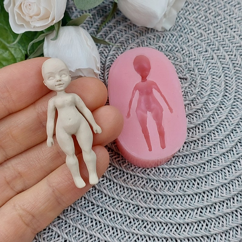 Op de afbeelding: Een roze siliconen mal voor het maken van een poppenfiguur. De mal is gevormd als een persoon met armen, benen en een hoofd. Naast de mal is een klein, wit poppenfiguur te zien.