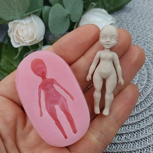 Op de afbeelding: Een roze siliconen mal voor het maken van een poppenfiguur. De mal is gevormd als een persoon met een hoofd, armen, benen en torso. De mal is gevormd als een persoon met een hoofd, armen, benen en torso. Naast de mal is een klein, ongeverfd poppenfiguur te zien.