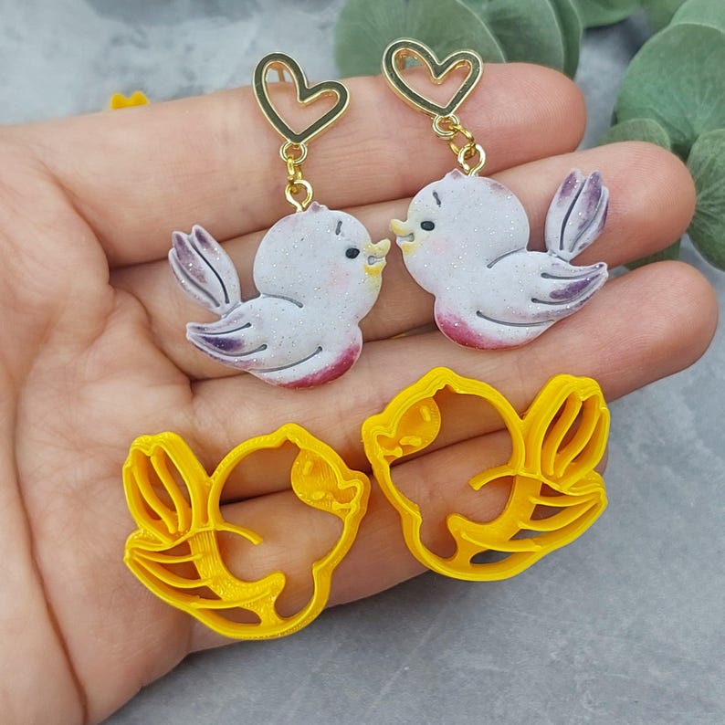 May include: A pair of earrings featuring two white birds with pink accents, kissing. The birds are attached to gold heart-shaped charms with a gold chain. The earrings are displayed on a hand with two yellow bird-shaped cookie cutters.