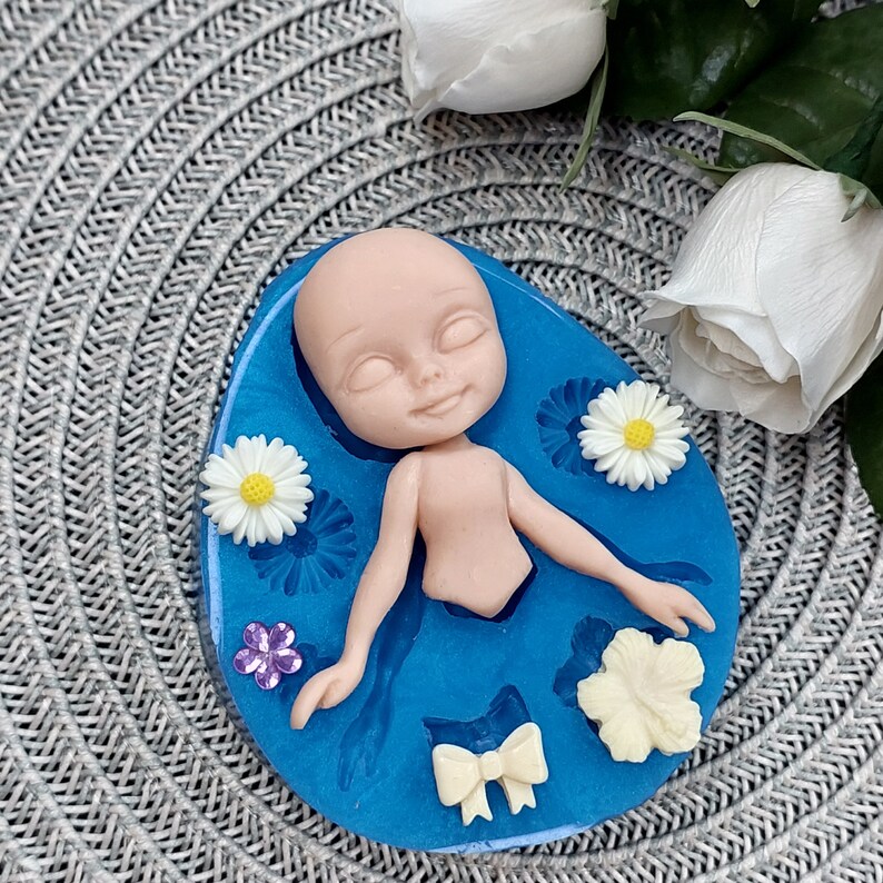 Pode incluir: Um molde de silicone azul com uma cavidade em forma de boneca e v&aacute;rias cavidades em forma de flores e la&ccedil;os. O molde est&aacute; sobre uma superf&iacute;cie cinza texturizada.