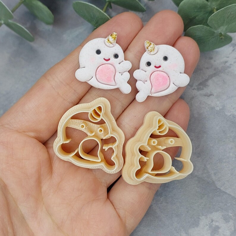 May include: Two white narwhal cookie cutters with gold glitter horns and pink accents. Two beige narwhal cookie cutters with gold glitter horns and pink accents.