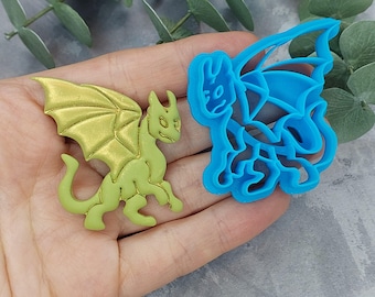 Könnte beinhalten: Eine grüne Drachenfigur mit goldenen Flügeln und ein blaues Drachen-Ausstecher. Die Drachenfigur ist detailliert mit einem strukturierten Körper und Flügeln. Der Ausstecher ist eine vereinfachte Umrisszeichnung eines Drachen.