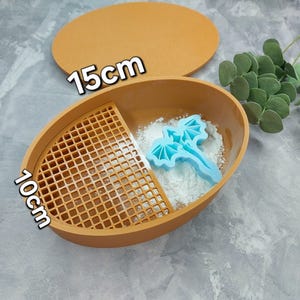 Puede incluir: Un recipiente ovalado marrón con tapa, que mide 15 cm de diámetro y 10 cm de alto. El recipiente tiene un inserto de malla y un cortador de galletas con forma de dragón de plástico azul en su interior. El recipiente está lleno de polvo blanco.