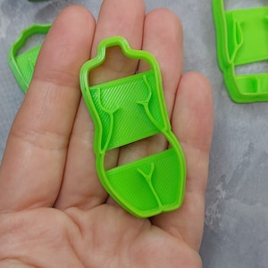 May include: Bright green plastic cookie cutter in the shape of a torso. The cutter has a rectangular midsection with diagonal lines and a detailed outline of the body. The cutter is held in a hand.