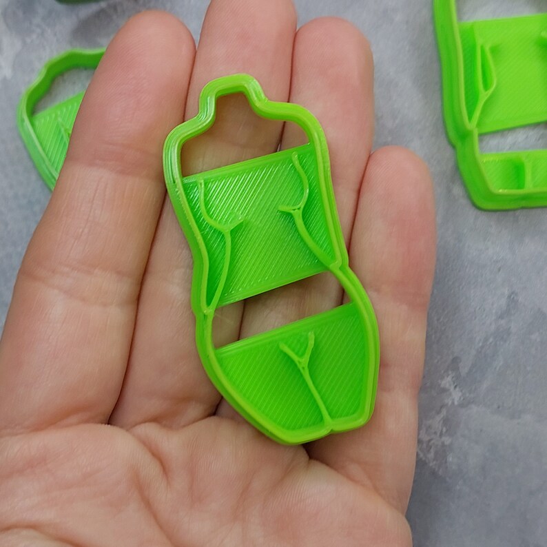 May include: Bright green plastic cookie cutter in the shape of a torso. The cutter has a rectangular midsection with diagonal lines and a detailed outline of the body. The cutter is held in a hand.