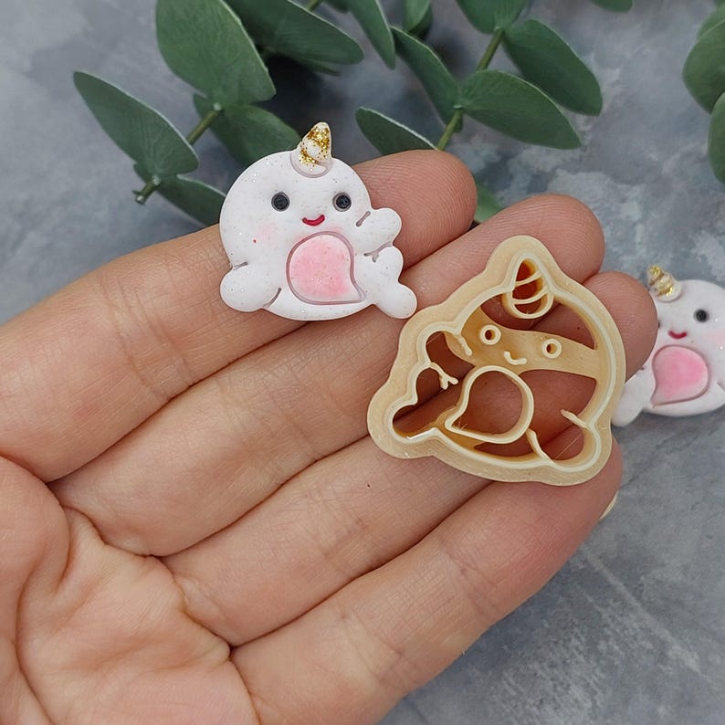 May include: Two cookie cutters, one in the shape of a narwhal and the other in the shape of a narwhal holding a heart. The narwhal cookie cutter is beige and the narwhal holding a heart cookie cutter is white.