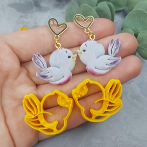 May include: A pair of earrings featuring a white bird with pink accents and a gold heart charm. The earrings are shown next to a pair of yellow bird-shaped cookie cutters.