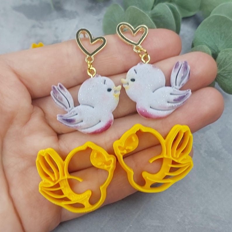 May include: A pair of earrings featuring a white bird with pink accents and a gold heart charm. The earrings are shown next to a pair of yellow bird-shaped cookie cutters.