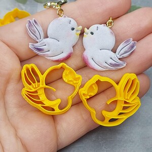 May include: Two pairs of 3D printed cookie cutters, one pair shaped like birds and the other pair shaped like flowers. The bird cutters are white with purple accents and the flower cutters are yellow.