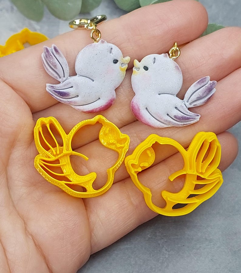 May include: Two pairs of 3D printed cookie cutters, one pair shaped like birds and the other pair shaped like flowers. The bird cutters are white with purple accents and the flower cutters are yellow.