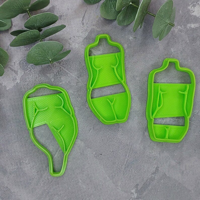 May include: Three bright green cookie cutters in the shape of a human body. The cutters are made of plastic and have a ribbed texture. The cutters are arranged on a gray surface with green leaves in the background.