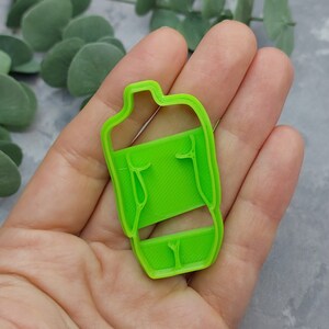 May include: A bright green plastic cookie cutter in the shape of a person's torso. The cutter is held in a hand, and the design includes the outline of the body with details like the shoulders and legs. The cutter is made of a solid, bright green plastic.