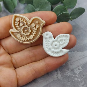 May include: Two dove-shaped cookie cutters. One is beige, the other is white. Both have floral and leaf patterns inside the dove shapes. The beige cutter is raised, while the white one is flat. The cutters are held in a hand.