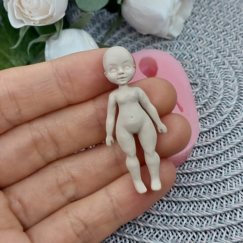 Op de afbeelding: Een klein, ongeverfd, wit poppenfiguurtje met een eenvoudig ontwerp. De pop staat op een roze siliconen mal.