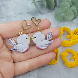 May include: A pair of earrings featuring a white bird with purple accents and a gold heart charm. The birds are facing each other and appear to be kissing.