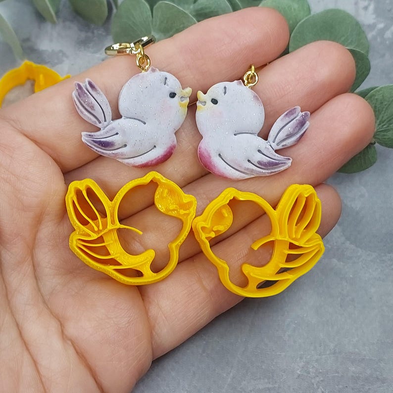 May include: Two pairs of earrings, one pair is a set of white bird earrings with gold accents, the other pair is a set of yellow bird-shaped earrings.