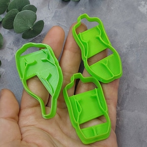 May include: Three bright green plastic cookie cutters in the shape of a leaf and two water bottles. The cookie cutters are held in a hand against a gray background. The leaf cutter has a detailed vein pattern.