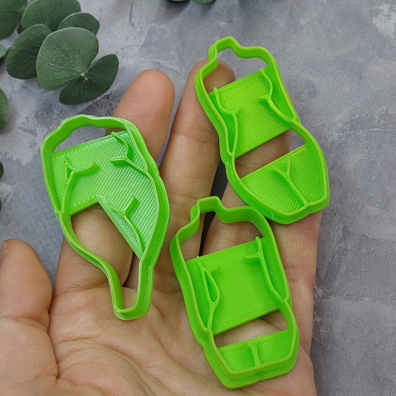 May include: Three bright green plastic cookie cutters in the shape of a leaf and two water bottles. The cookie cutters are held in a hand against a gray background. The leaf cutter has a detailed vein pattern.