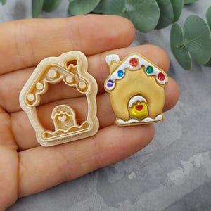 Puede incluir: Un cortador de galletas de casa de jengibre y una galleta de casa de jengibre decorada. El cortador de galletas es beige y tiene un corazón blanco en el centro. La galleta es beige con glaseado blanco y caramelos de colores.