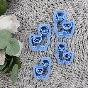May include: Four blue plastic cookie cutters shaped like llamas. Each llama has a decorative design on its neck.