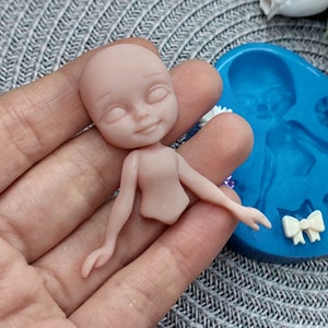 Pode incluir: Uma boneca de cor nude com os olhos fechados e um leve sorriso. A boneca &eacute; feita de argila polim&eacute;rica e est&aacute; sendo segurada em uma m&atilde;o. A boneca est&aacute; ao lado de um molde de silicone azul de uma boneca semelhante.