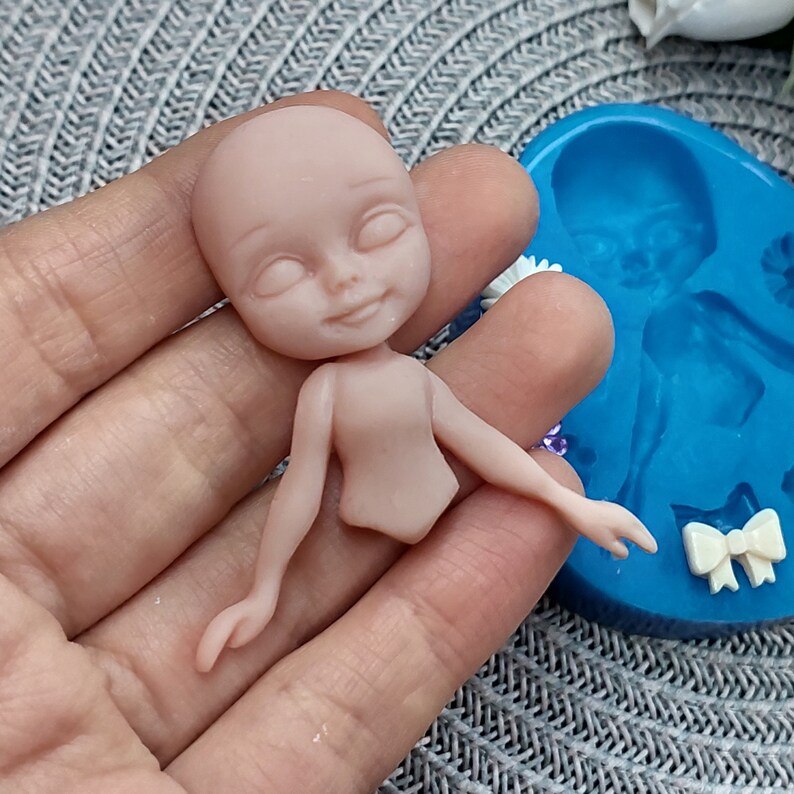 Pode incluir: Uma boneca de cor nude com os olhos fechados e um leve sorriso. A boneca &eacute; feita de argila polim&eacute;rica e est&aacute; sendo segurada em uma m&atilde;o. A boneca est&aacute; ao lado de um molde de silicone azul de uma boneca semelhante.