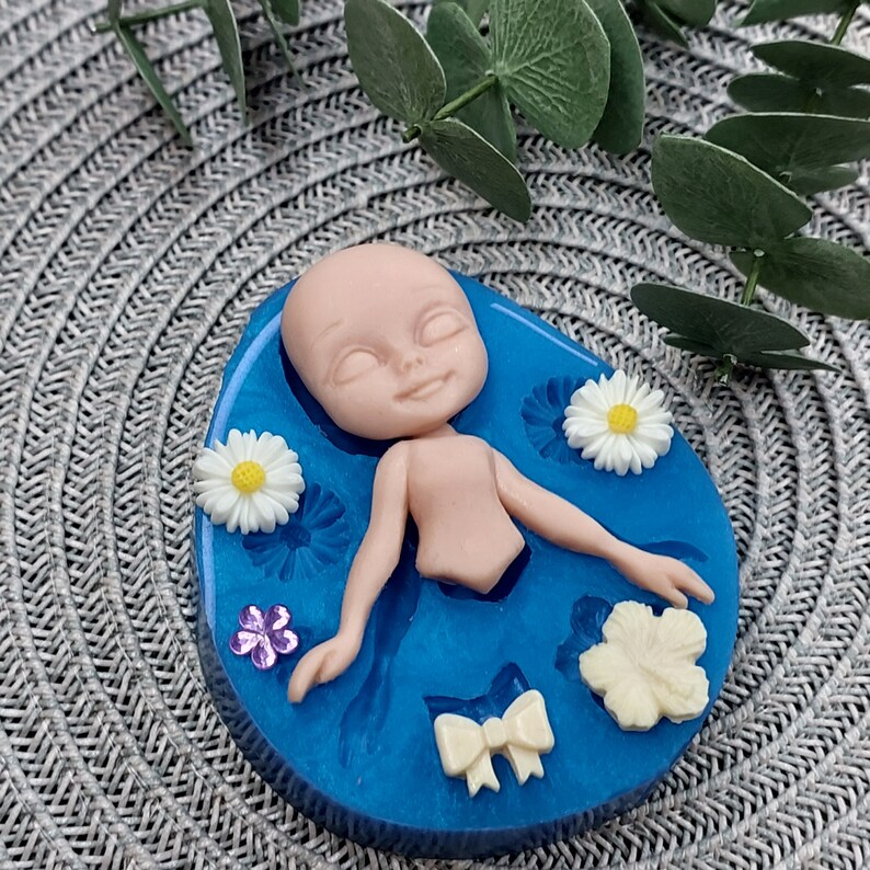 Pode incluir: Um molde de silicone azul com uma cavidade em forma de boneca, cercada por cavidades em forma de flores e la&ccedil;os. A boneca est&aacute; em uma pose relaxada com os olhos fechados.