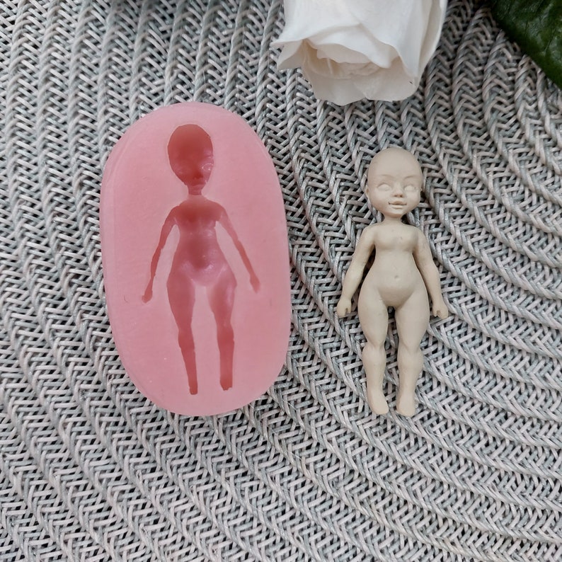 Op de afbeelding: Een roze siliconen mal voor het maken van een poppenfiguur. De mal is gevormd als een persoon met armen, benen en een hoofd. Naast de mal is een klein, ongeverfd poppenfiguur te zien.