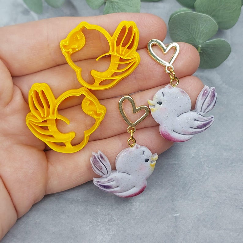 May include: Two pairs of earrings featuring a bird and heart design. The earrings are made of polymer clay and gold metal. The birds are painted in shades of purple and white, and the hearts are gold.