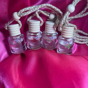 May include: Four small glass bottles with a floral design and the text "Doniqua Divine" on each bottle. The bottles are hanging from a white and gold rope with wooden beads.