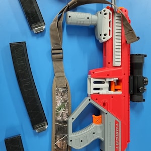 Op de afbeelding: Een rood en grijs speelgoedpistool met een camouflageband, een richtkijker en een magazijn. Het pistool heeft de woorden "TOMCAT" en "MIRAX" erop gedrukt. Het pistool wordt ook geleverd met drie zwarte magazijnen en een grijze en oranje dart-houder.