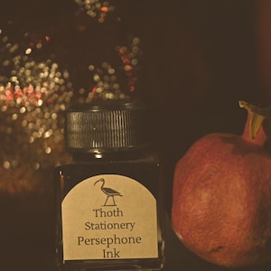May include: A bottle of "Persephone Ink" from "Thoth Stationery" with a black cap and a label featuring a bird illustration. A ripe pomegranate is next to the ink bottle. The background has bokeh lights.
