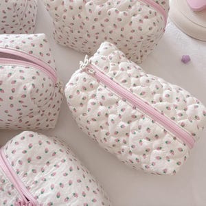 May include: A collection of white quilted cosmetic bags, patterned with pink strawberries and pink zippers. The bags are rectangular and have a quilted texture. The zippers are open and closed. The bags are arranged on a white surface.