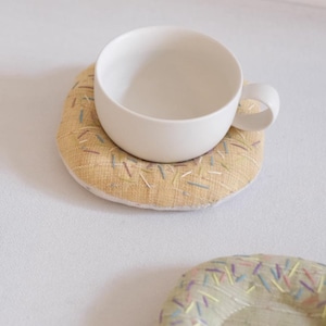 Może przedstawiać: Biały ceramiczny kubek do herbaty spoczywa na podkładce w kształcie pączka. Podkładka jest żółta z haftowanymi posypkami w różnych kolorach. Widoczna jest kolejna podkładka.