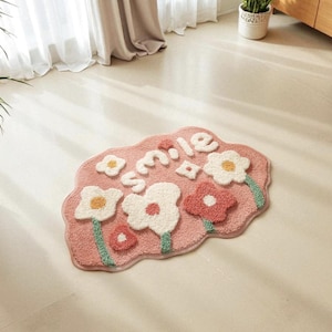 Puede incluir: Una alfombra rosa con forma de flor con la palabra "smile" en letras blancas. La alfombra presenta varias flores grandes, blancas y rojas con tallos verdes. La alfombra está hecha de un material suave y texturizado.