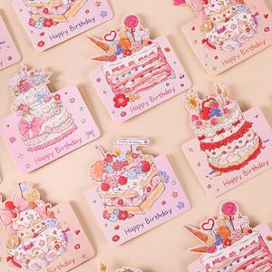 May include: A selection of pink birthday cards featuring illustrated layered cakes decorated with flowers, bows, and sweets. Each card is printed with the words "Happy Birthday".