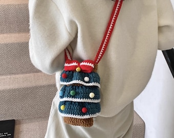 May include: A handmade crochet crossbody bag shaped like a Christmas tree. The bag is primarily teal with brown and white accents, and colorful pom-poms. It has a red strap and a red bow at the top. The bag is worn over the shoulder.