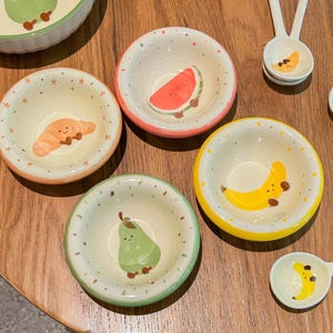 May include: A collection of small, hand-painted ceramic bowls with fruit and pastry designs. The bowls feature a watermelon, banana, pear, and croissant. Each bowl has a different colored rim, and the background is speckled with small dots.