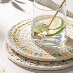 May include: A clear glass filled with water, a lime slice, and a gold spoon rests on a white, circular coaster. The coaster is decorated with yellow and green floral embroidery. Another coaster is stacked below.