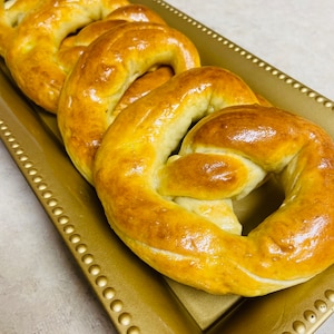 May include: A close-up of four golden brown pretzels on a gold tray. The pretzels are soft and shiny, with a slightly glossy finish.