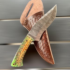 May include: A Damascus steel knife with a patterned blade and a handle featuring green, yellow, and brown tones. The knife is paired with a brown leather sheath with embossed detailing. The knife is designed for outdoor use.