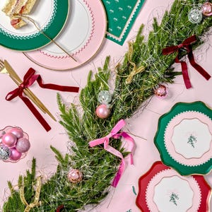 Puede incluir: Una mesa de comedor festiva con platos de papel verde y rosa, servilletas y una rebanada de pastel. La mesa est&aacute; decorada con una guirnalda de ramas de pino, cinta dorada y adornos. Las servilletas tienen un patr&oacute;n verde y rosa con la palabra "HOPE" impresa en ellas.