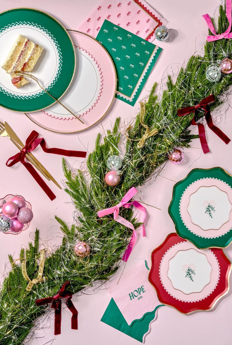 Puede incluir: Una mesa de comedor festiva con platos de papel verde y rosa, servilletas y una rebanada de pastel. La mesa est&aacute; decorada con una guirnalda de ramas de pino, cinta dorada y adornos. Las servilletas tienen un patr&oacute;n verde y rosa con la palabra "HOPE" impresa en ellas.