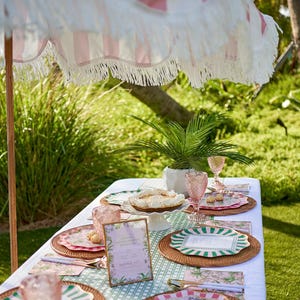 A Palm Beach inspired tablesetting with a green and white rattan inspired table runner. There are pink & green striped paper plates and matching cocktail napkins. There's a pink and white striped cabana umbrella. 
Framed menu that says Palm Royale Bachelorette Party