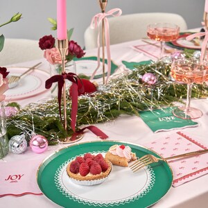 Puede incluir: Un plato de papel verde y blanco con un borde de encaje. El plato tiene un borde dorado y est&aacute; decorado con una tarta de frambuesas y una crema de hojaldre. El plato est&aacute; sobre un mantel rosa con una servilleta rosa y blanca que dice "JOY".