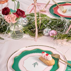 Puede incluir: Un plato de papel verde y rosa con un borde dorado y un centro blanco con un dise&ntilde;o de rama de pino verde. Un peque&ntilde;o pastel con glaseado rosa y chispas rojas est&aacute; en el plato.