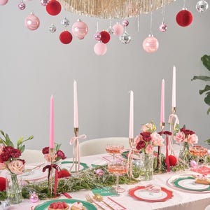 Puede incluir: Un mantel rosa con un plato estampado en verde y blanco, un plato estampado en rosa y rojo y una copa de champ&aacute;n rosado. Hay velas, flores y una guirnalda de oropel dorado y adornos rojos y rosas colgando sobre la mesa.